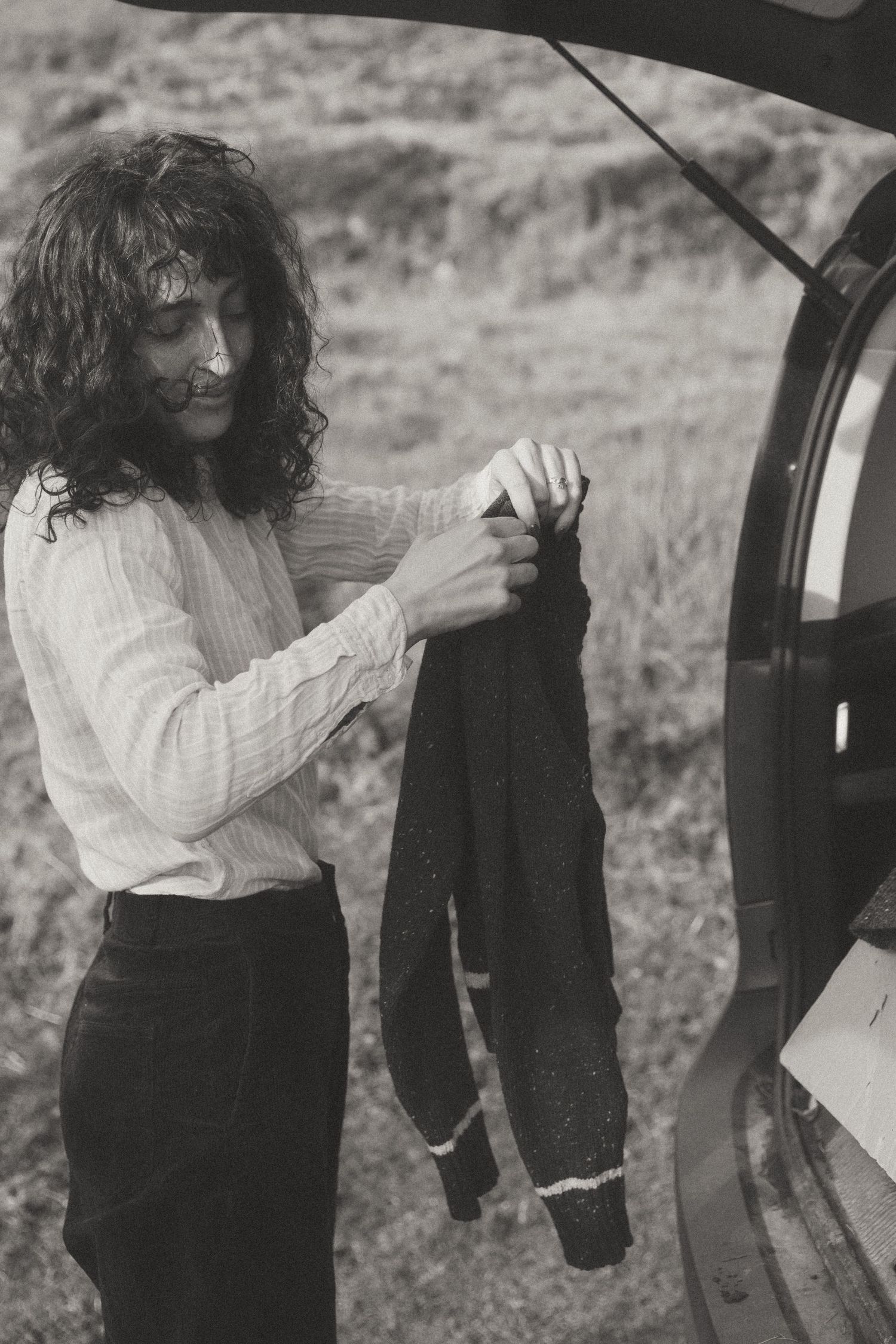 A person with long dark curly hair wearing a white shirt holds up dark clothing next to a car in a black and white photo.