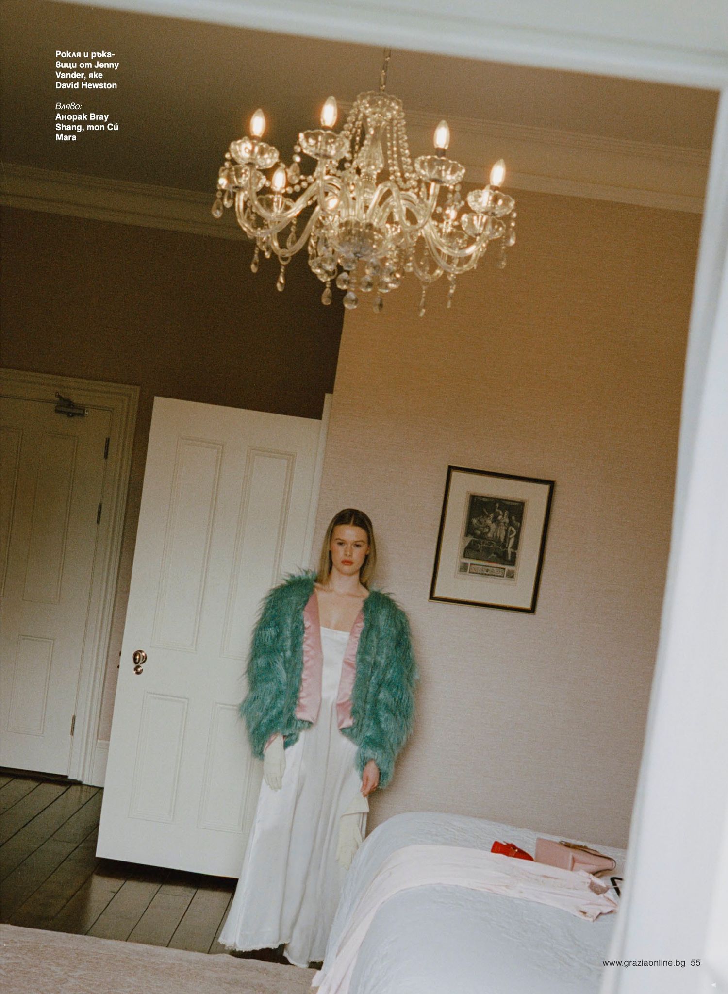 Woman in teal feather jacket and white trousers stands beneath crystal chandelier in elegant room.