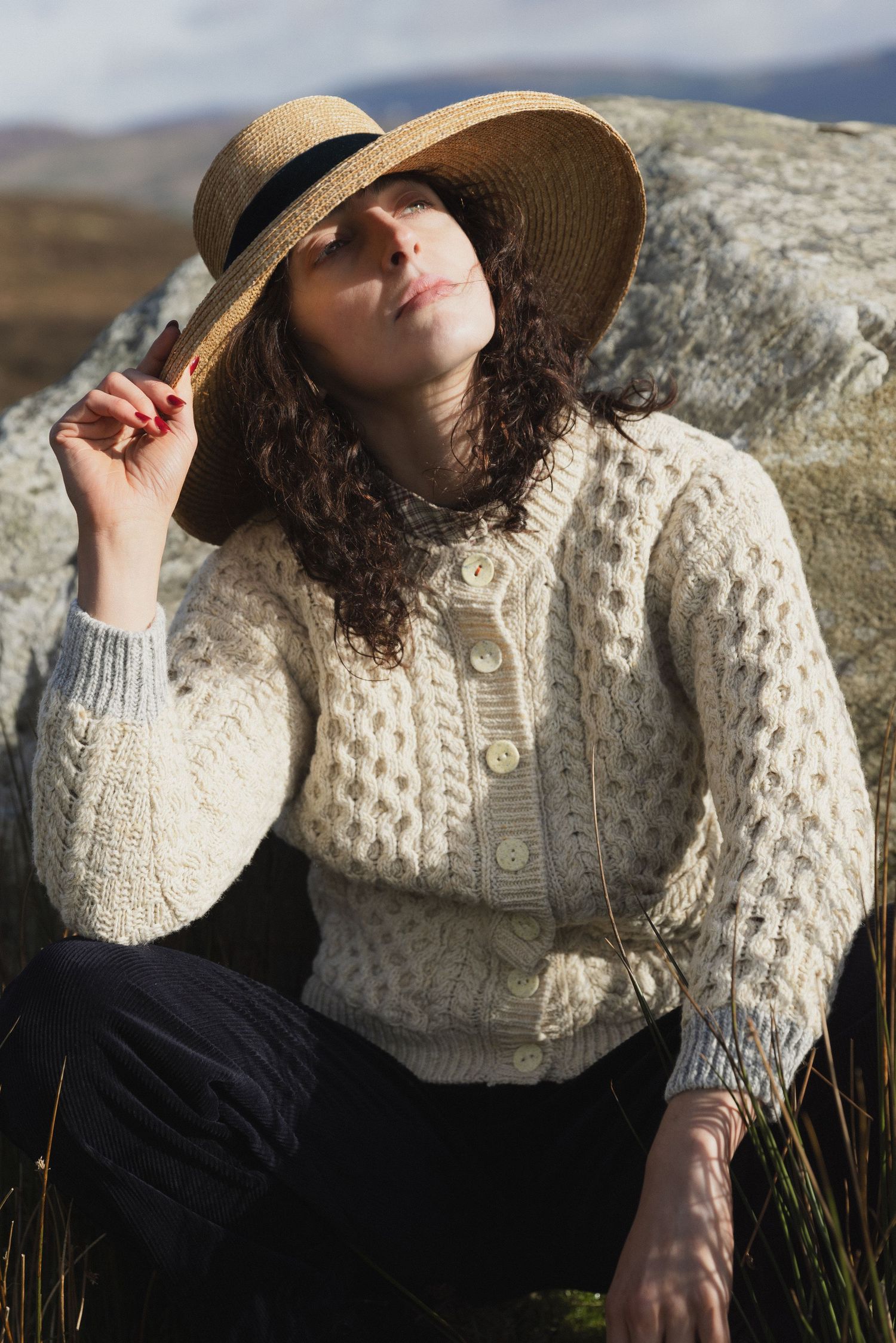 Person in cream cable knit sweater and straw hat lounges on rocks outdoors in natural sunlight.
