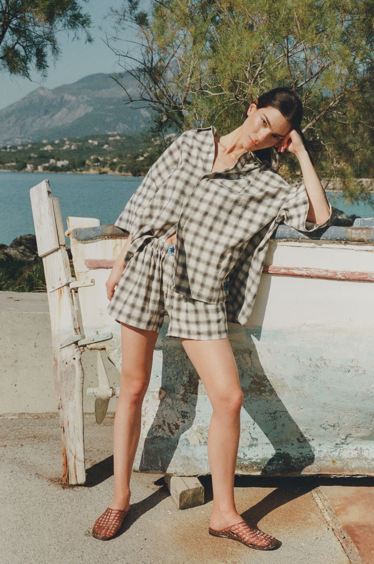 Woman in a matching gingham shirt and shorts set with woven flat sandals poses by a weathered boat on a seaside shore with mountains behind.
