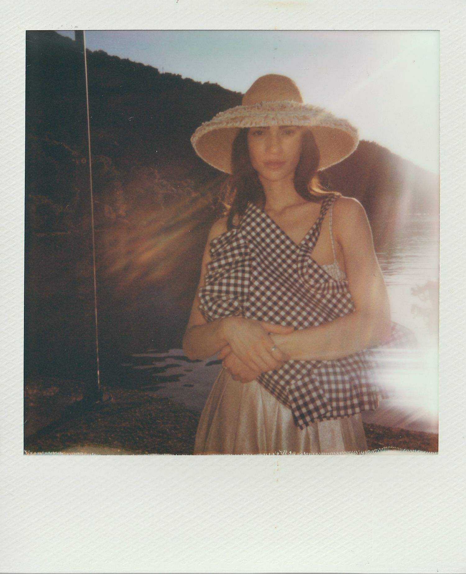 Polaroid-style photo of a woman in a wide-brim straw hat and black-and-white gingham ruffle top standing by a calm lake at sunset.