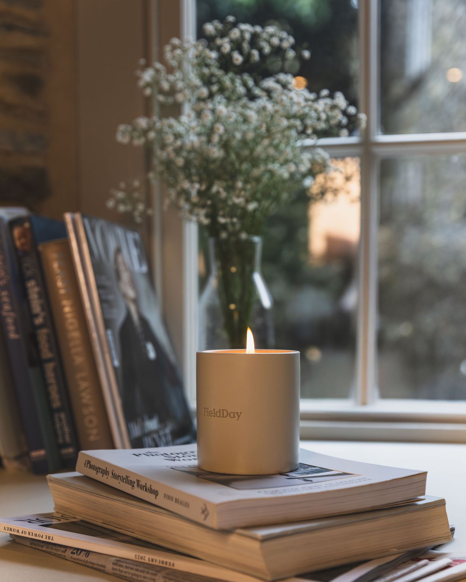 Stack of books with a beige candle on top near a window with soft natural lighting and white flowers in the background.