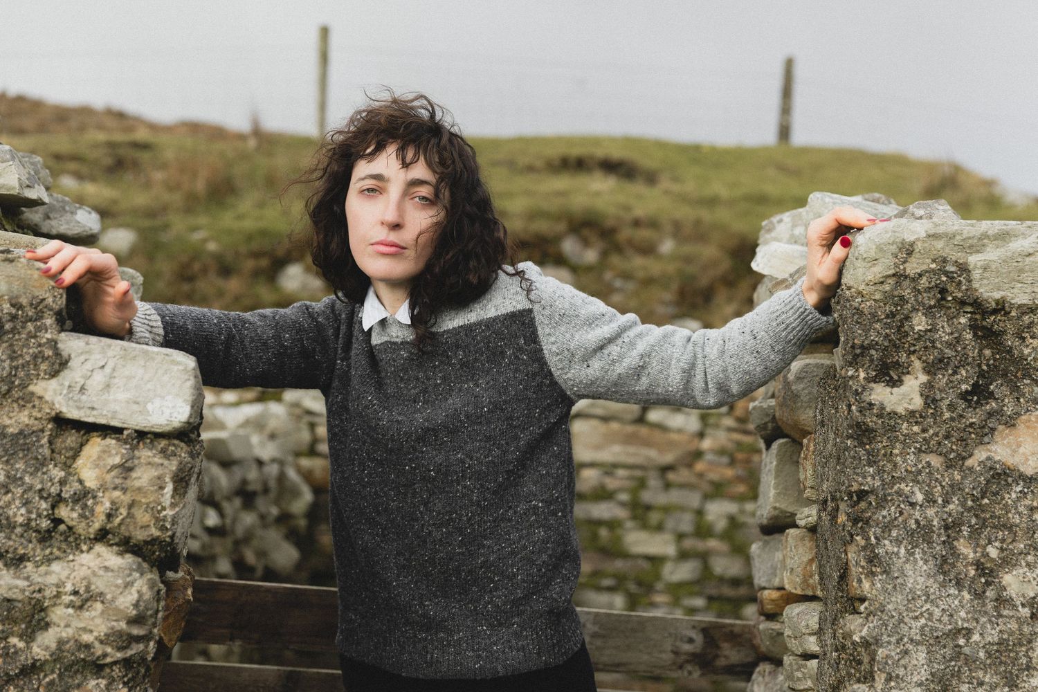 Person wearing a gray sweater poses against stone walls in a rustic outdoor setting.