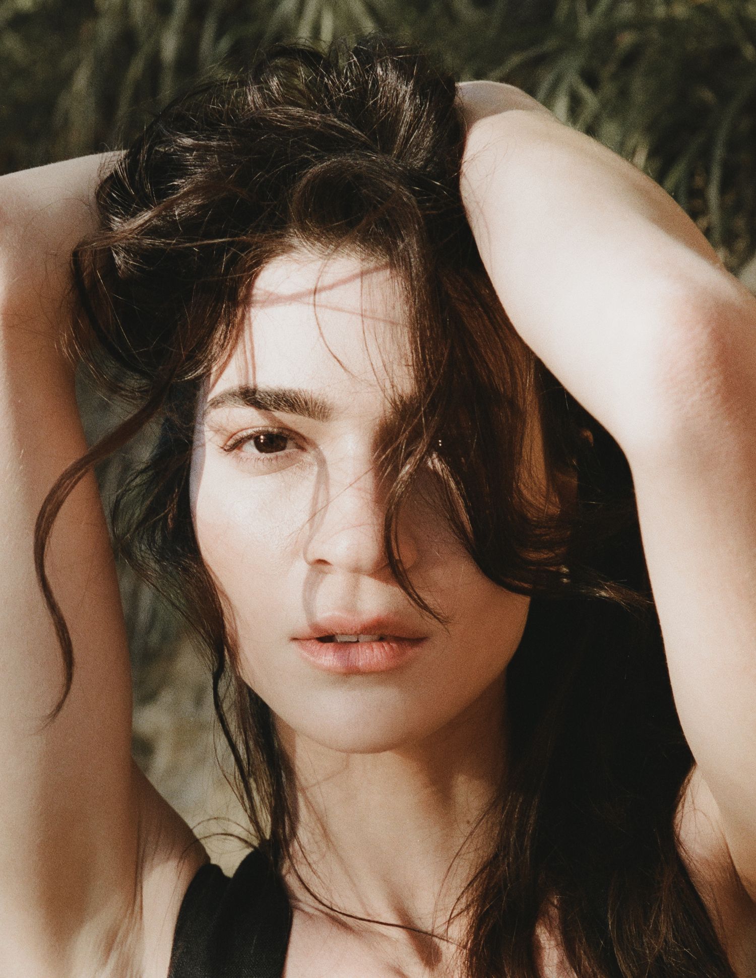 Woman with tousled dark hair and arms raised above her head poses outdoors in warm sunlight with green foliage behind her.