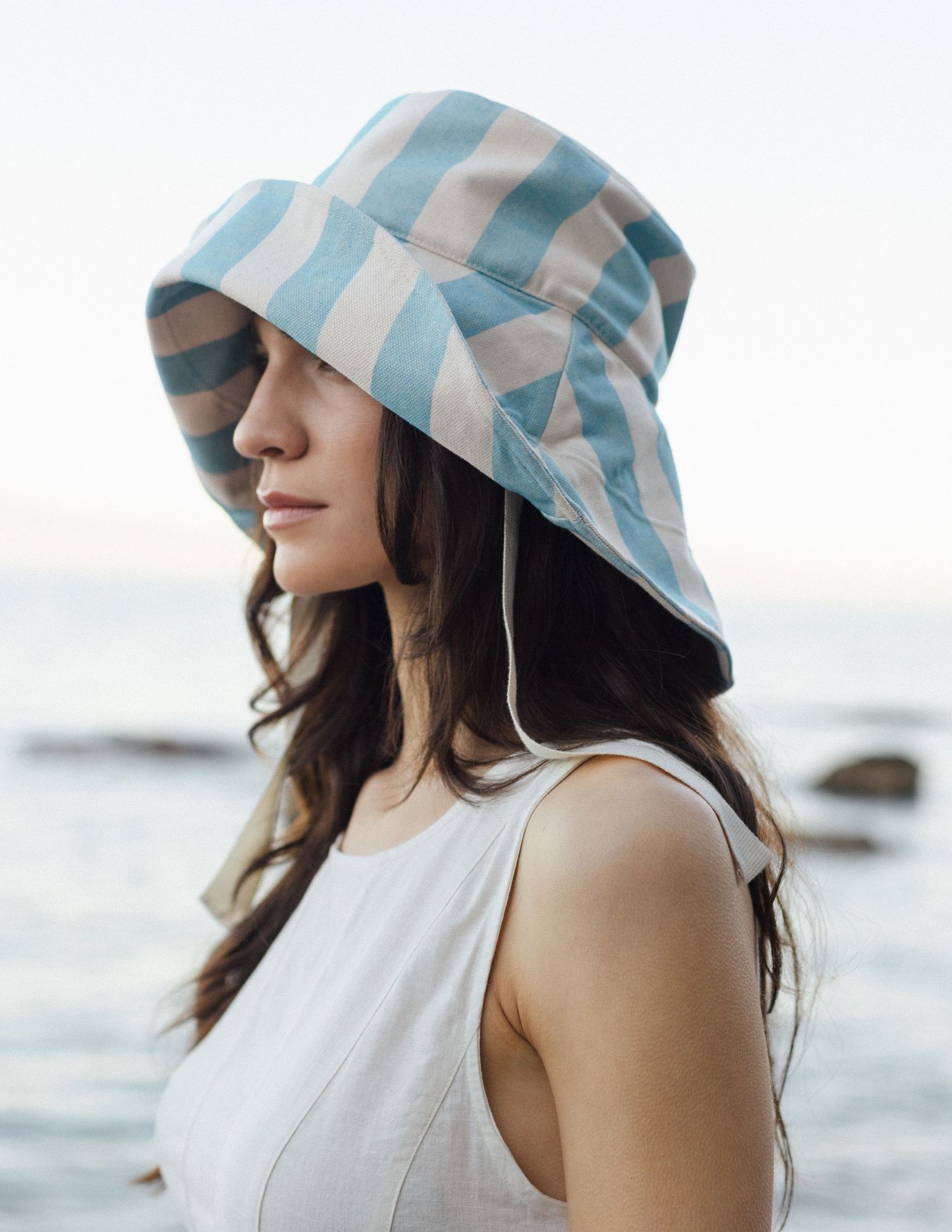 Woman in profile wearing a blue and cream striped bucket sun hat and white sleeveless top, standing by the ocean at soft sunset.