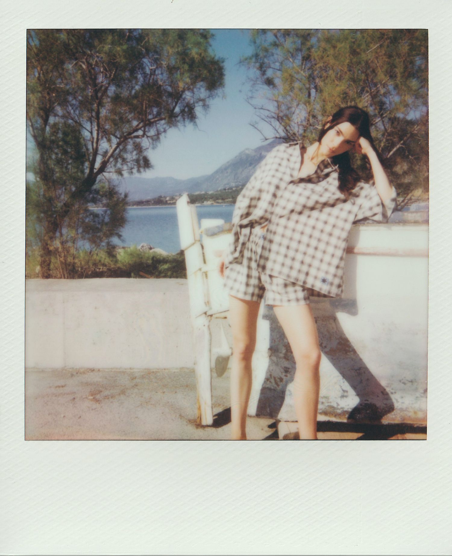 Polaroid of a woman in a plaid matching shirt and shorts leaning on a white wall beside a chair, with sea, mountains and trees behind.
