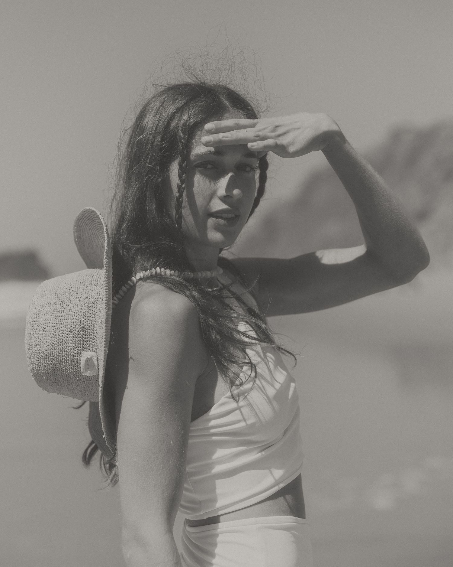 A person in a white top poses with a sun hat against a bright outdoor backdrop in a black and white photograph.