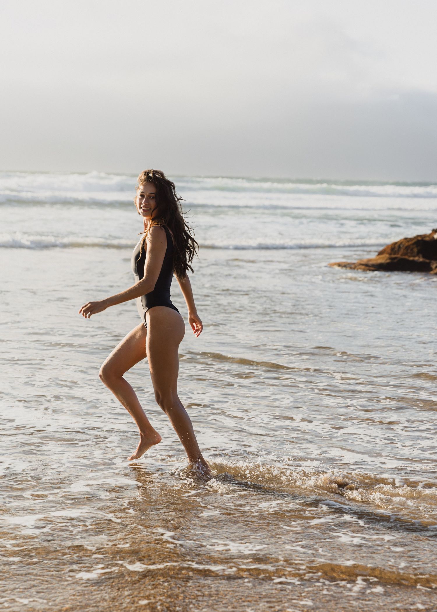Someone in a black swimsuit splashes and plays in the shallow ocean waves on a cloudy beach day.