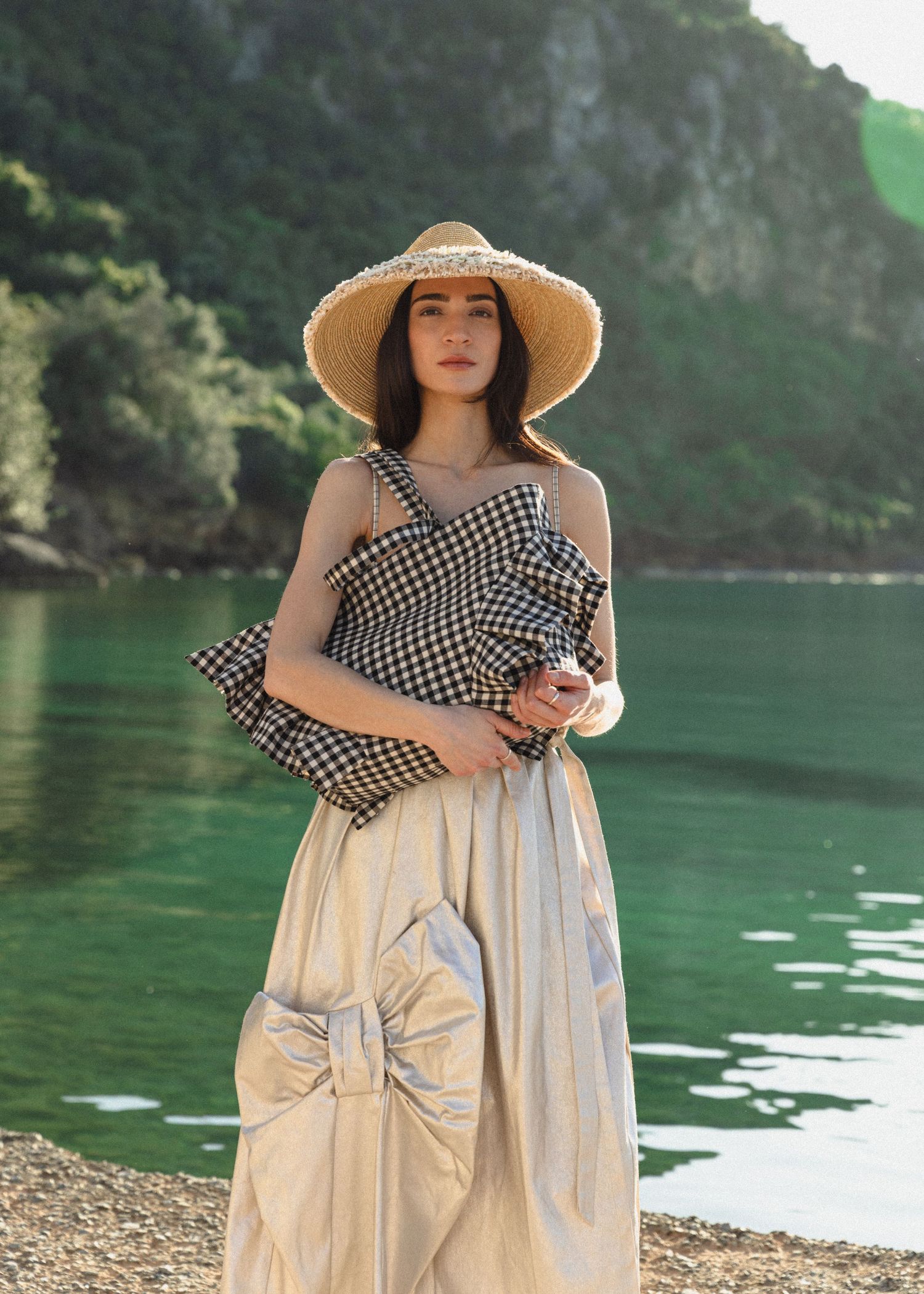 Woman in a straw hat, gingham top and cream bow-detail skirt stands on a pebble shore beside calm green water.