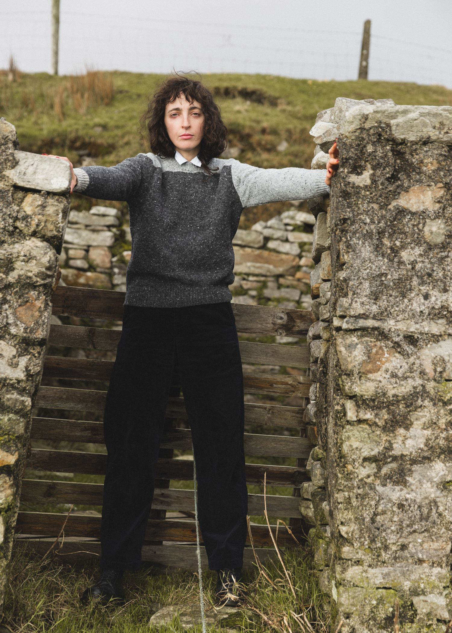 A person wearing a gray sweater and black pants stands against a rustic stone wall in a natural outdoor setting.