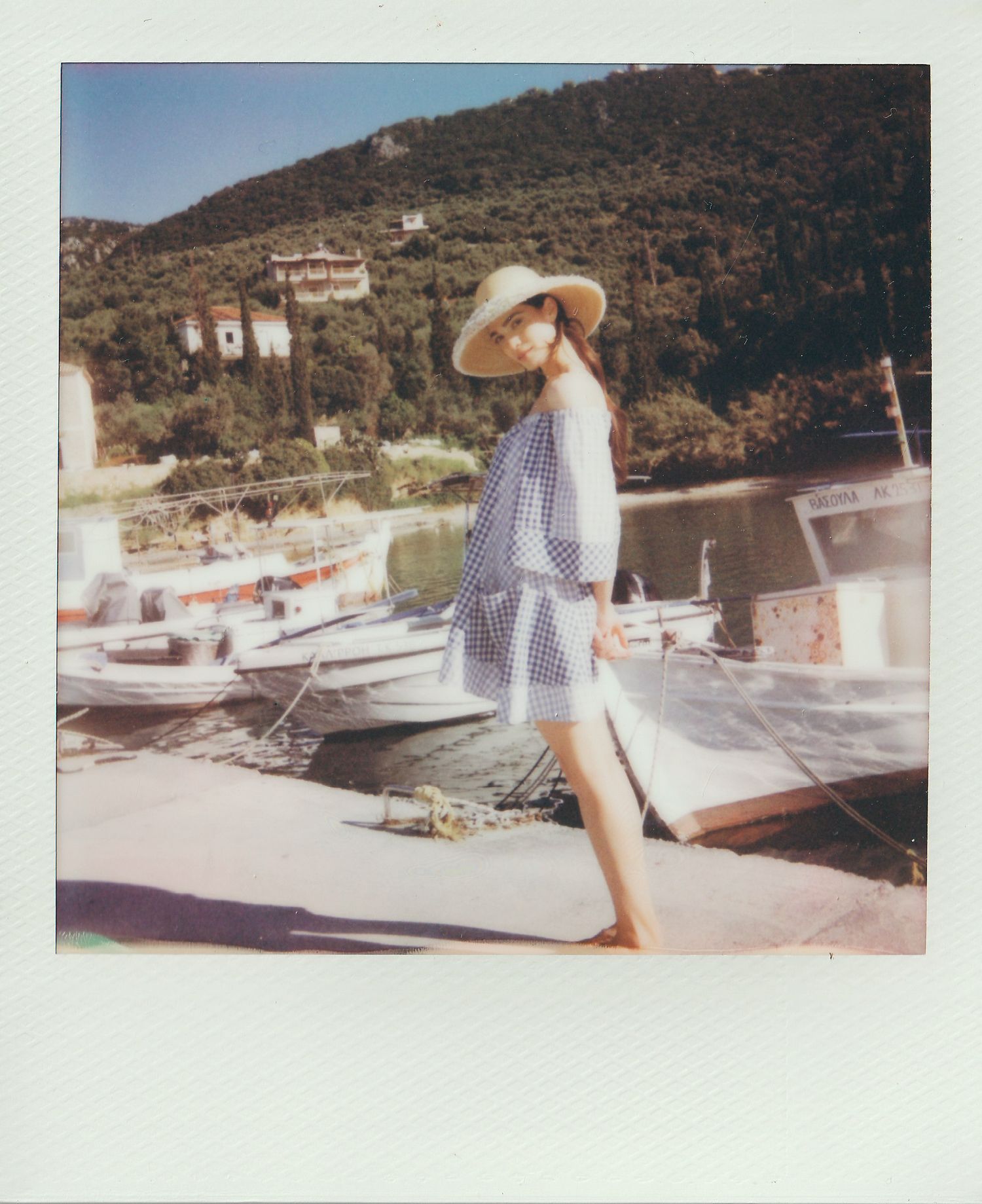 Woman in a straw hat and blue gingham dress stands on a harbor dock beside moored fishing boats, with green hills behind in a Polaroid.