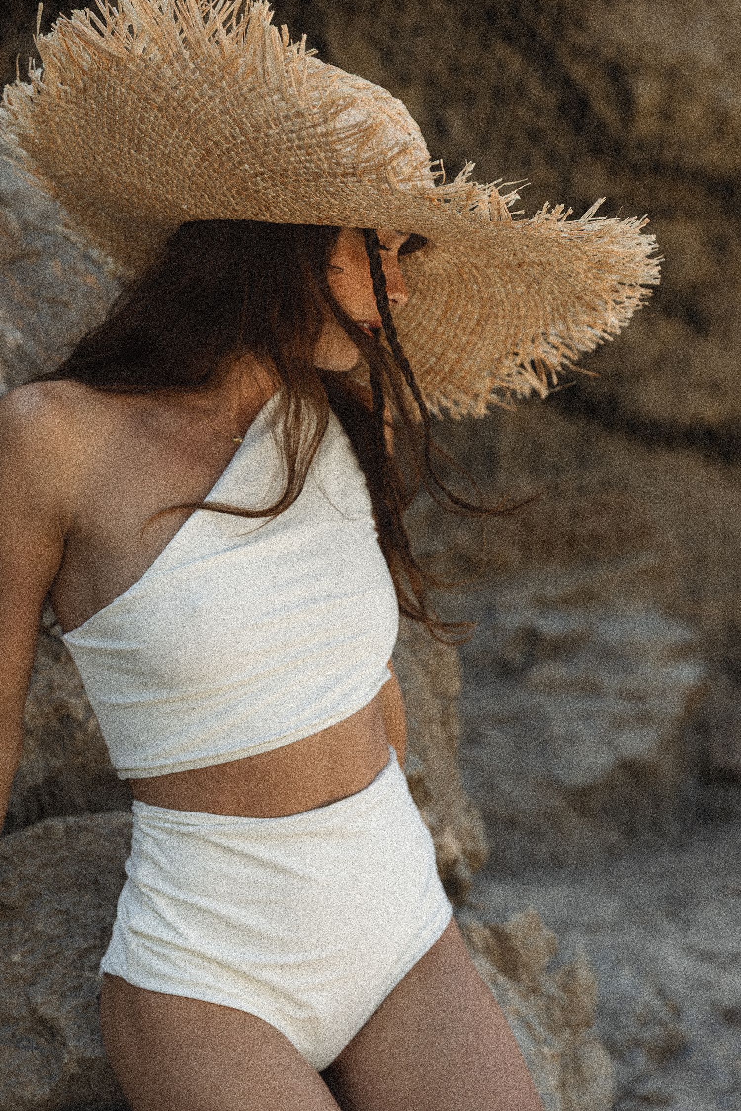 Woman in white two-piece swimsuit and frayed straw hat poses against rocky backdrop in summer beach fashion campaign shot.