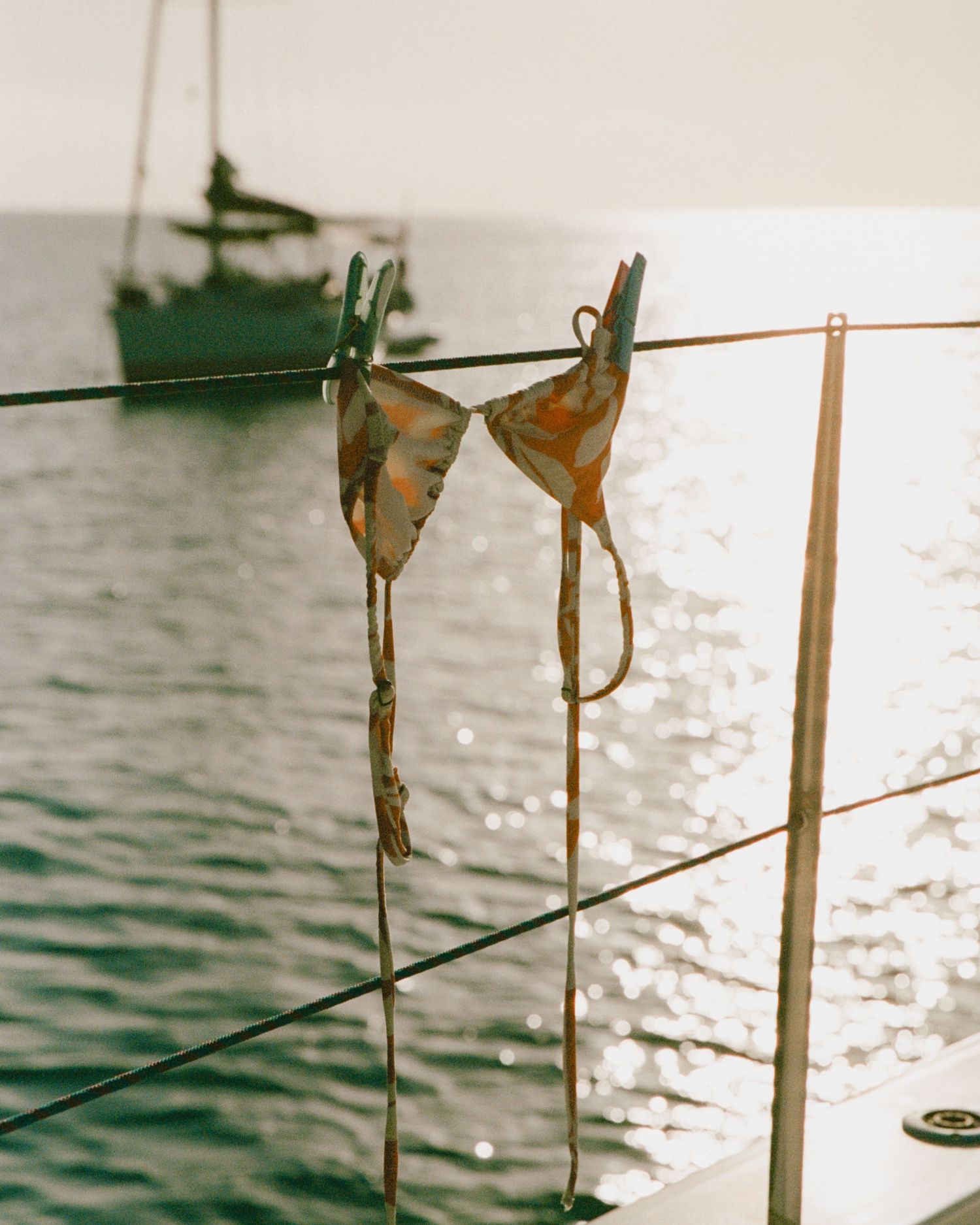 A floral bikini top hangs to dry on a boat railing at sunset, with a sailboat anchored on the sparkling ocean behind.