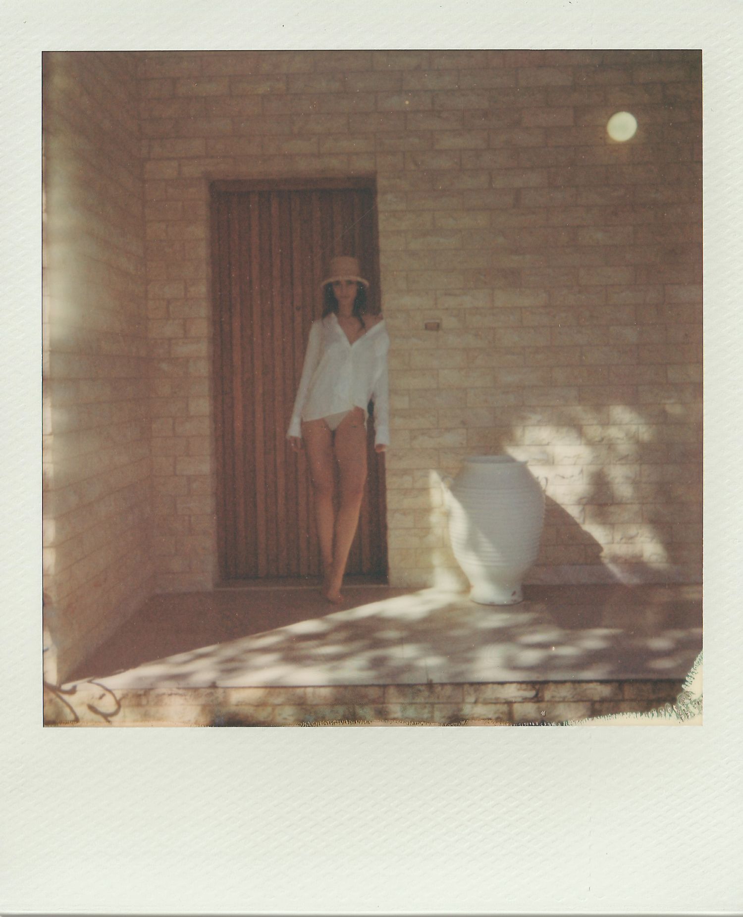 Polaroid photo of a woman in a wide-brimmed hat and white shirt standing barefoot in a sunlit brick doorway beside a large white urn.