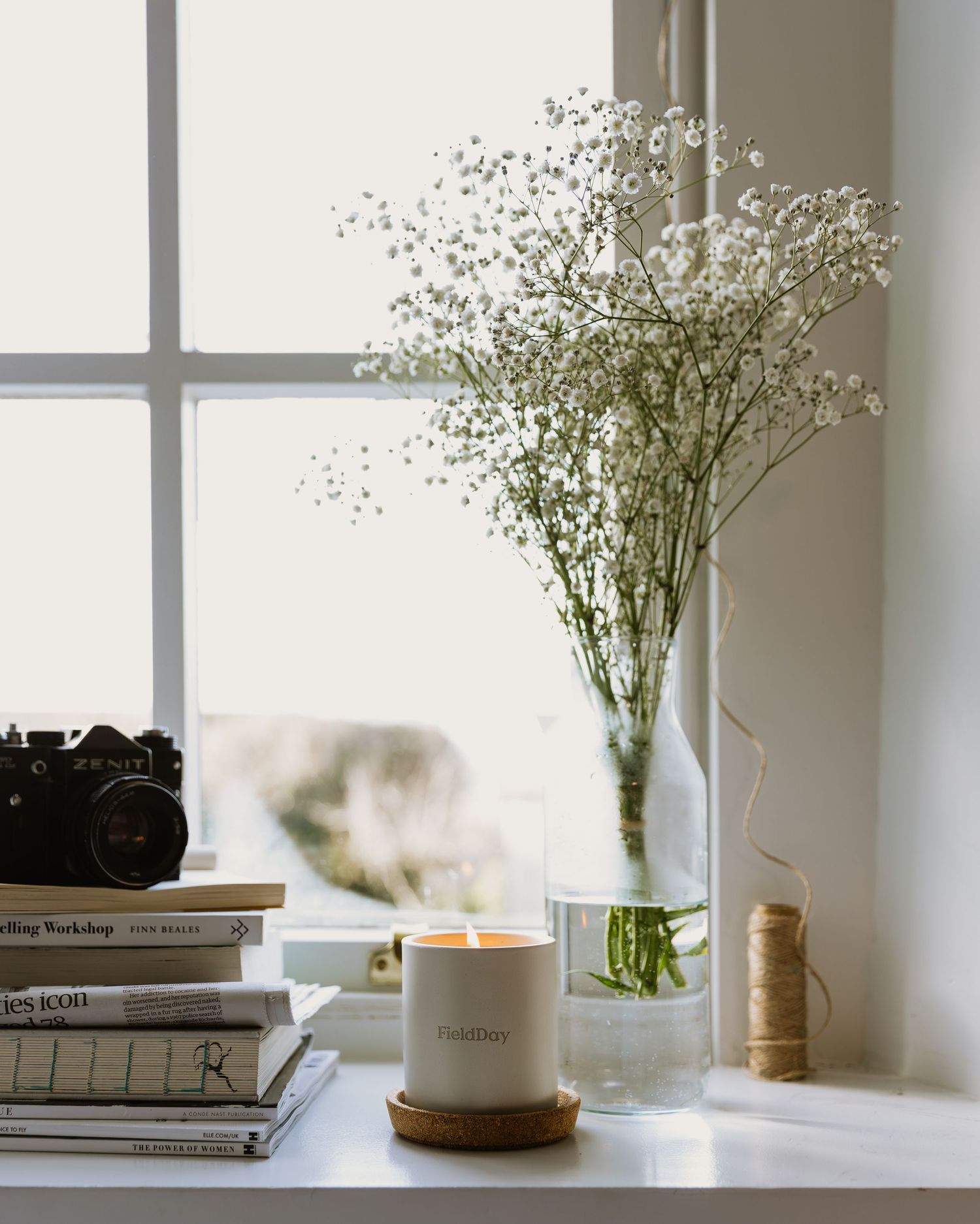 White baby's breath flowers in a glass vase sit on a windowsill with books and a candle in natural light.
