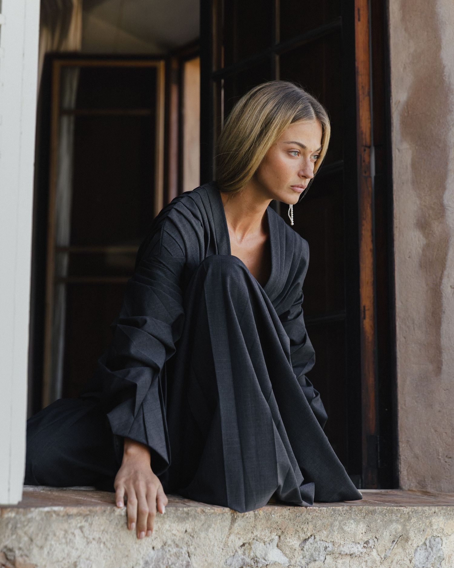 Woman with long blonde hair in a flowing black outfit sits on a weathered stone ledge, gazing to the side beside an open wooden window.
