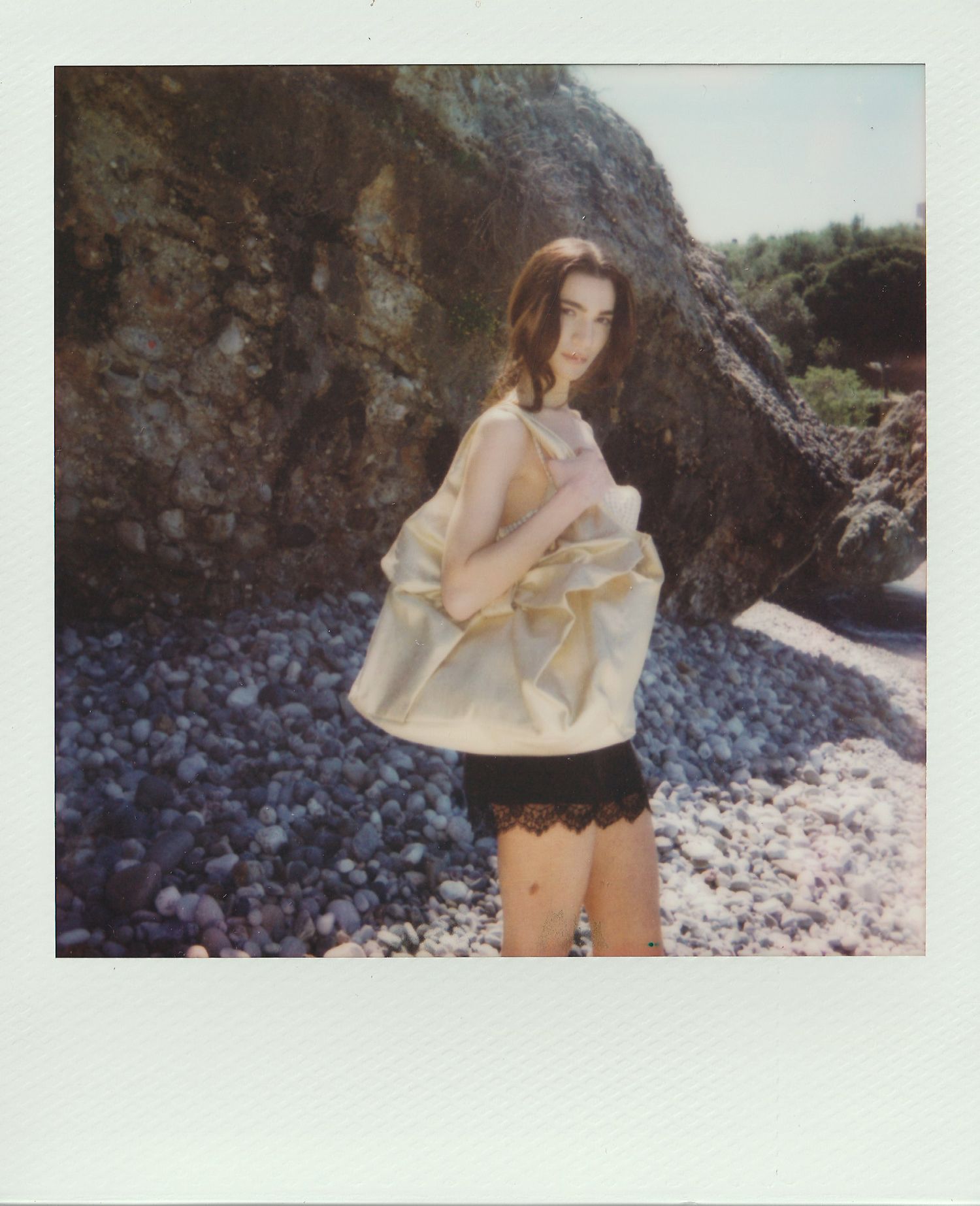 Polaroid-style photo of a woman in a black lace skirt holding a large metallic gold tote bag on a pebble beach beside a rocky cliff.
