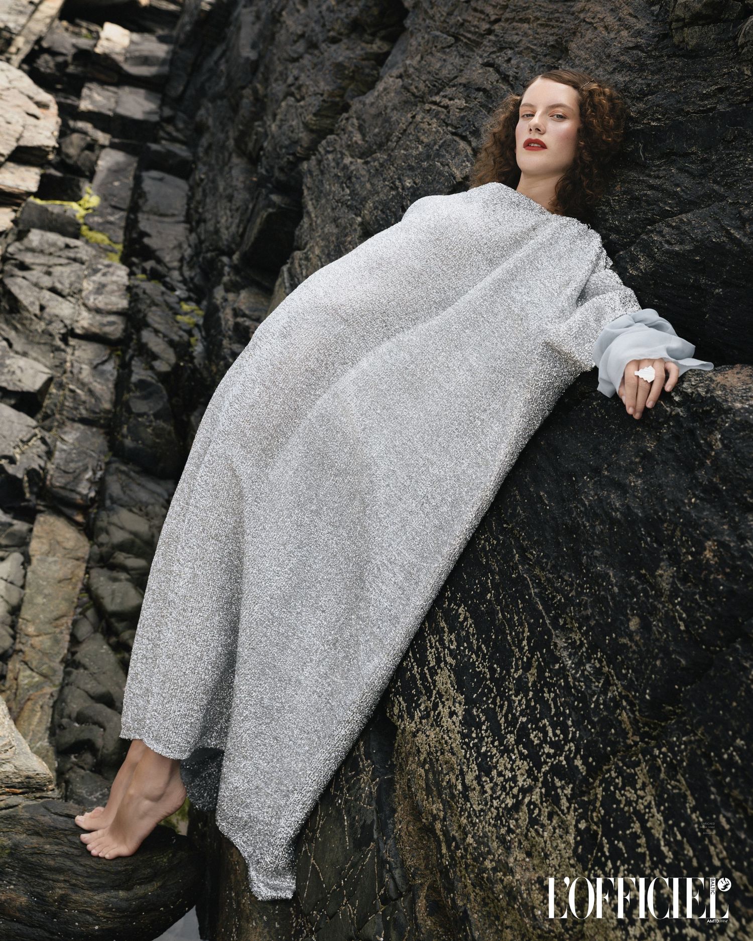 Fashion model in sparkly silver dress poses dramatically against dark rocky cliff face editorial shoot for L'officiel magazine.