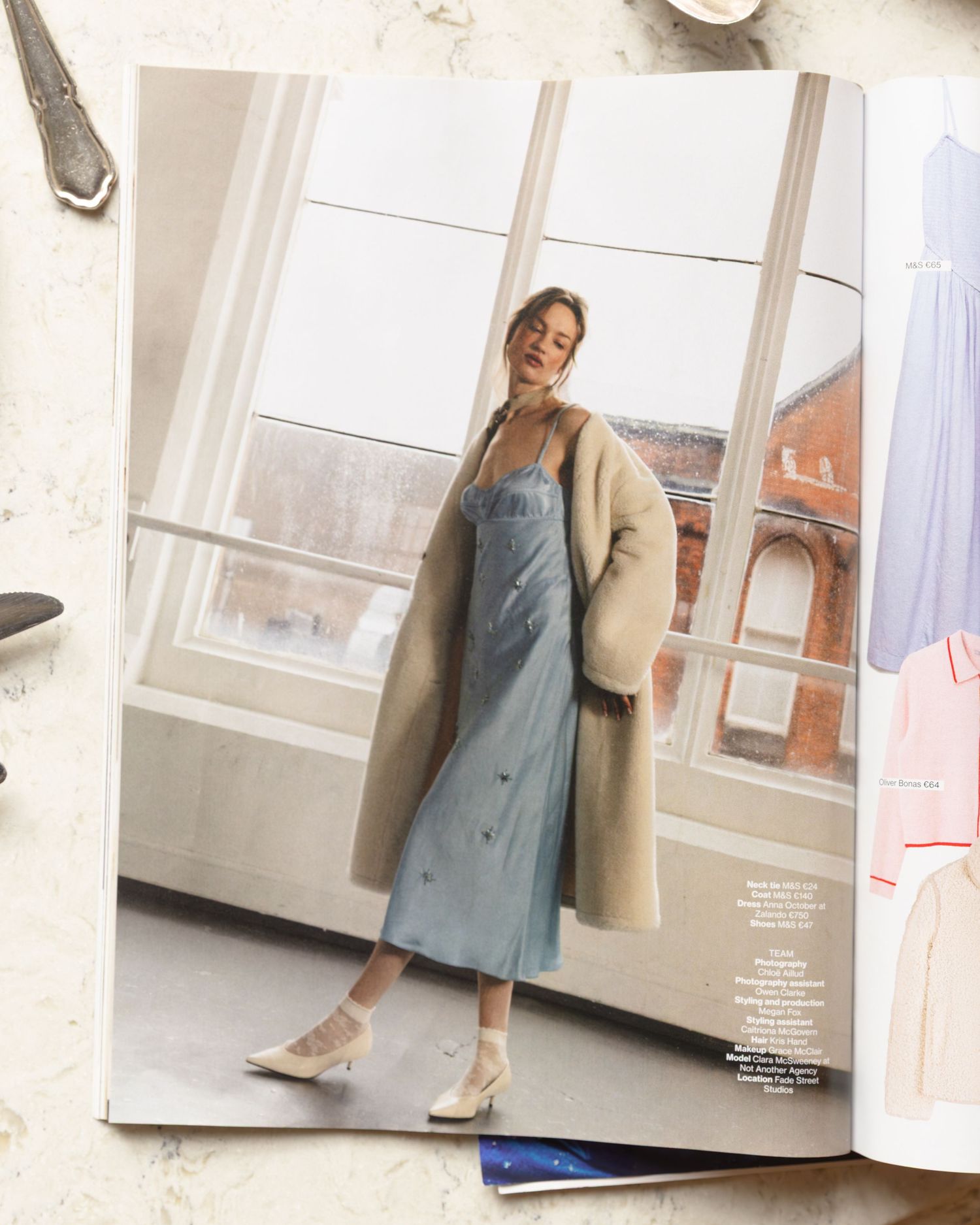 A model in a denim dress and beige coat poses elegantly in a bright minimalist studio setting.