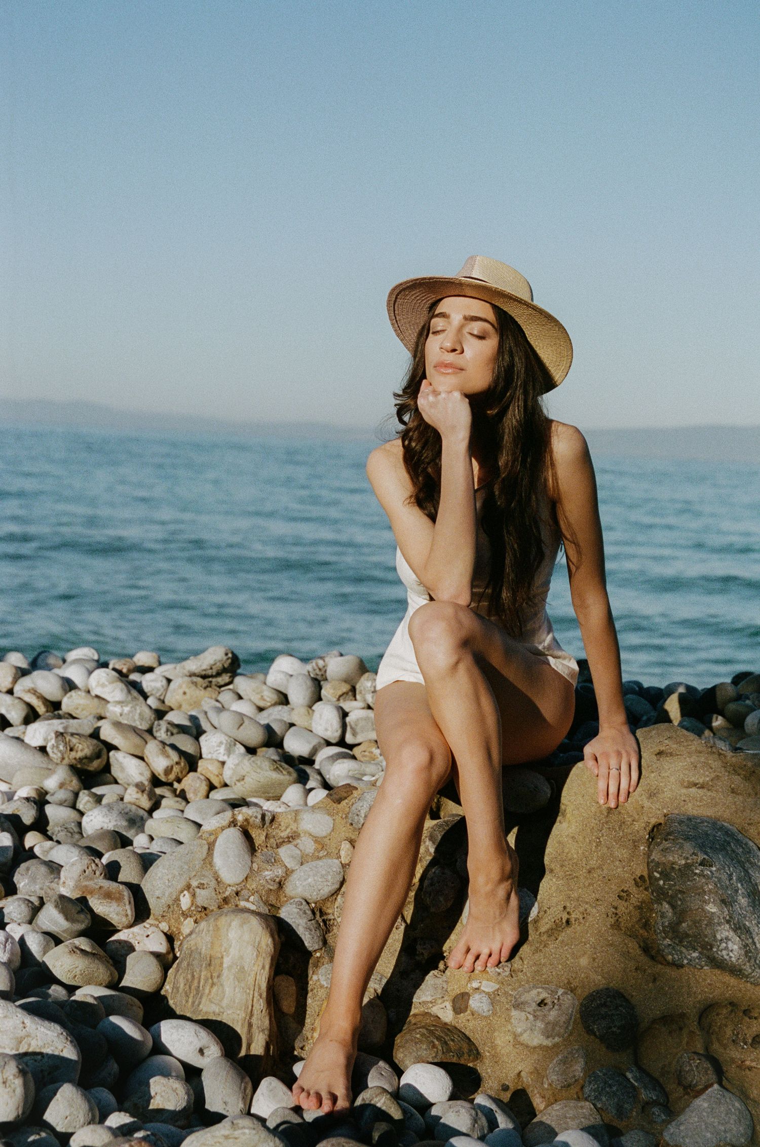Woman in a straw hat and white outfit sits barefoot on a large rock with eyes closed, surrounded by smooth pebbles and the calm blue ocean.