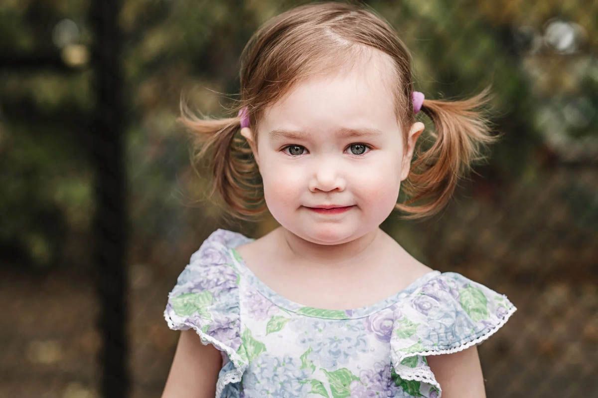 A child wearing a floral dress with white lace trim poses against a blurred outdoor background.