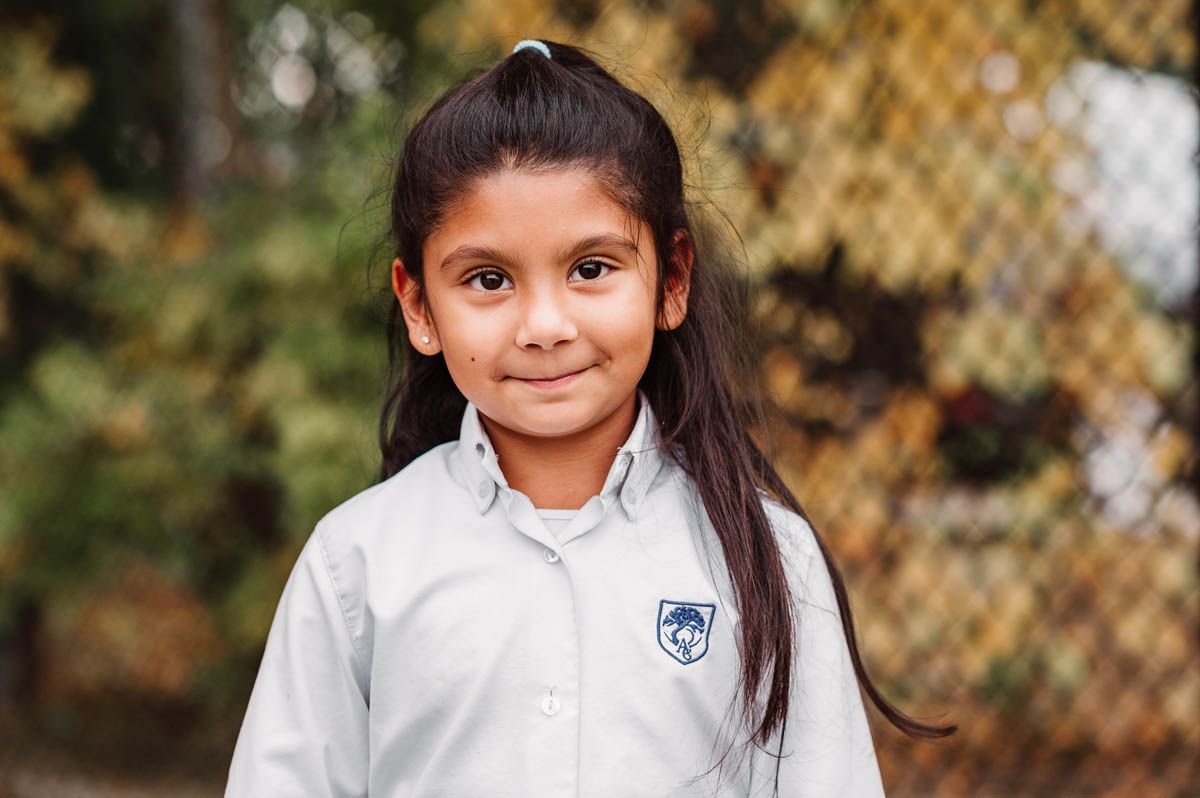 Student in white school uniform poses outdoors near chain link fence.