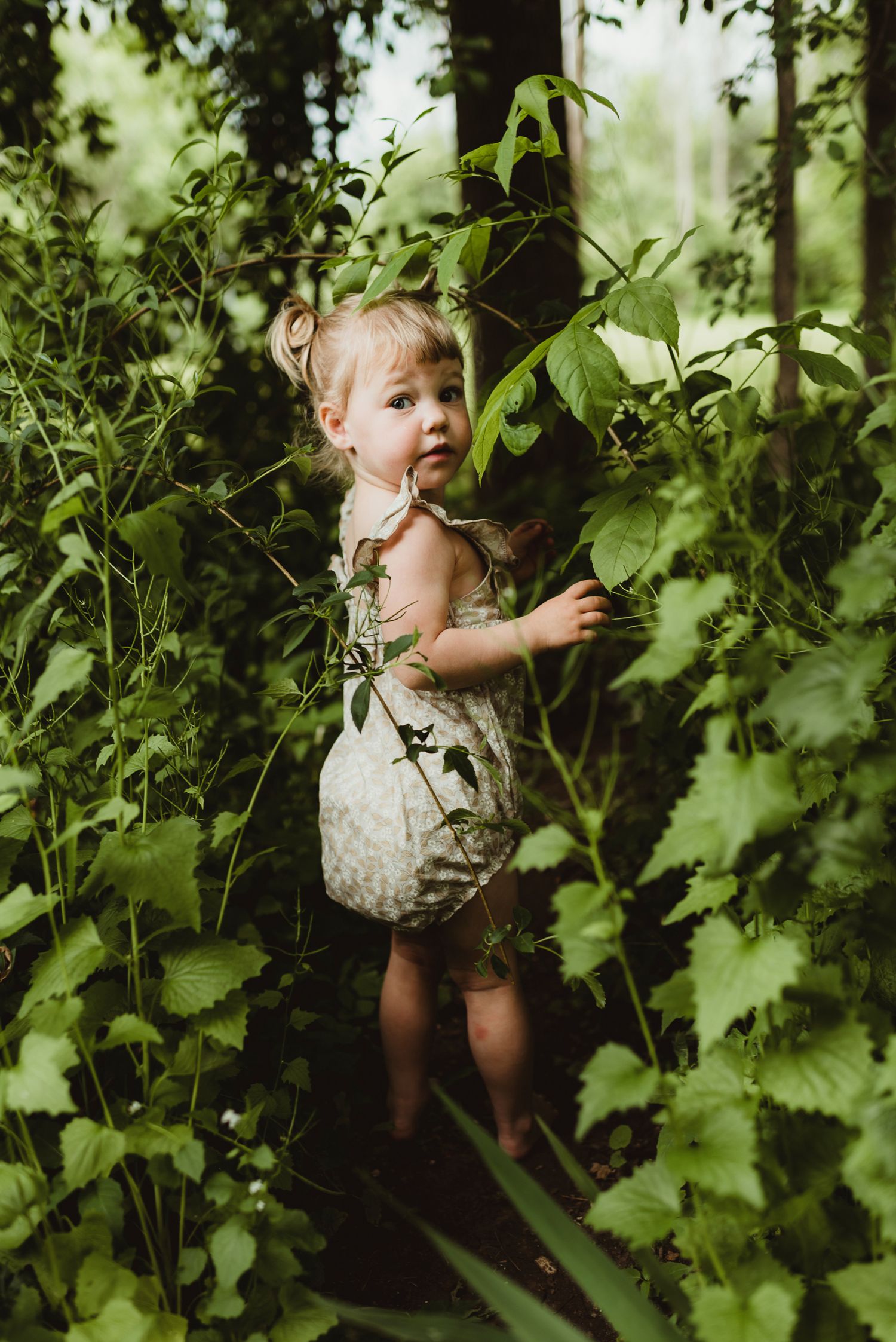 Small child in spotted dress exploring lush green foliage in a garden setting.