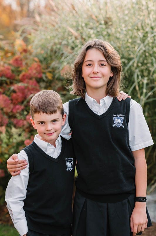 Two students in school uniforms pose together outdoors with white shirts and dark vests.