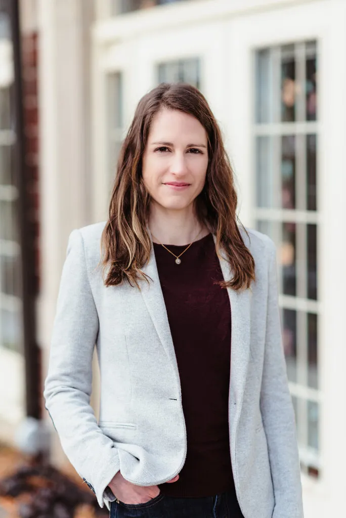 Professional businesswoman poses in light gray blazer and dark top against white building exterior.