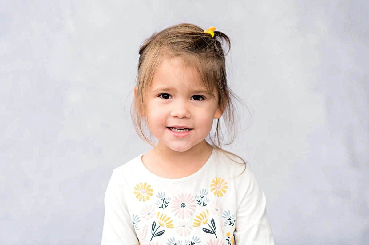 A child in a white dress smiles while playing in front of a light colored background.