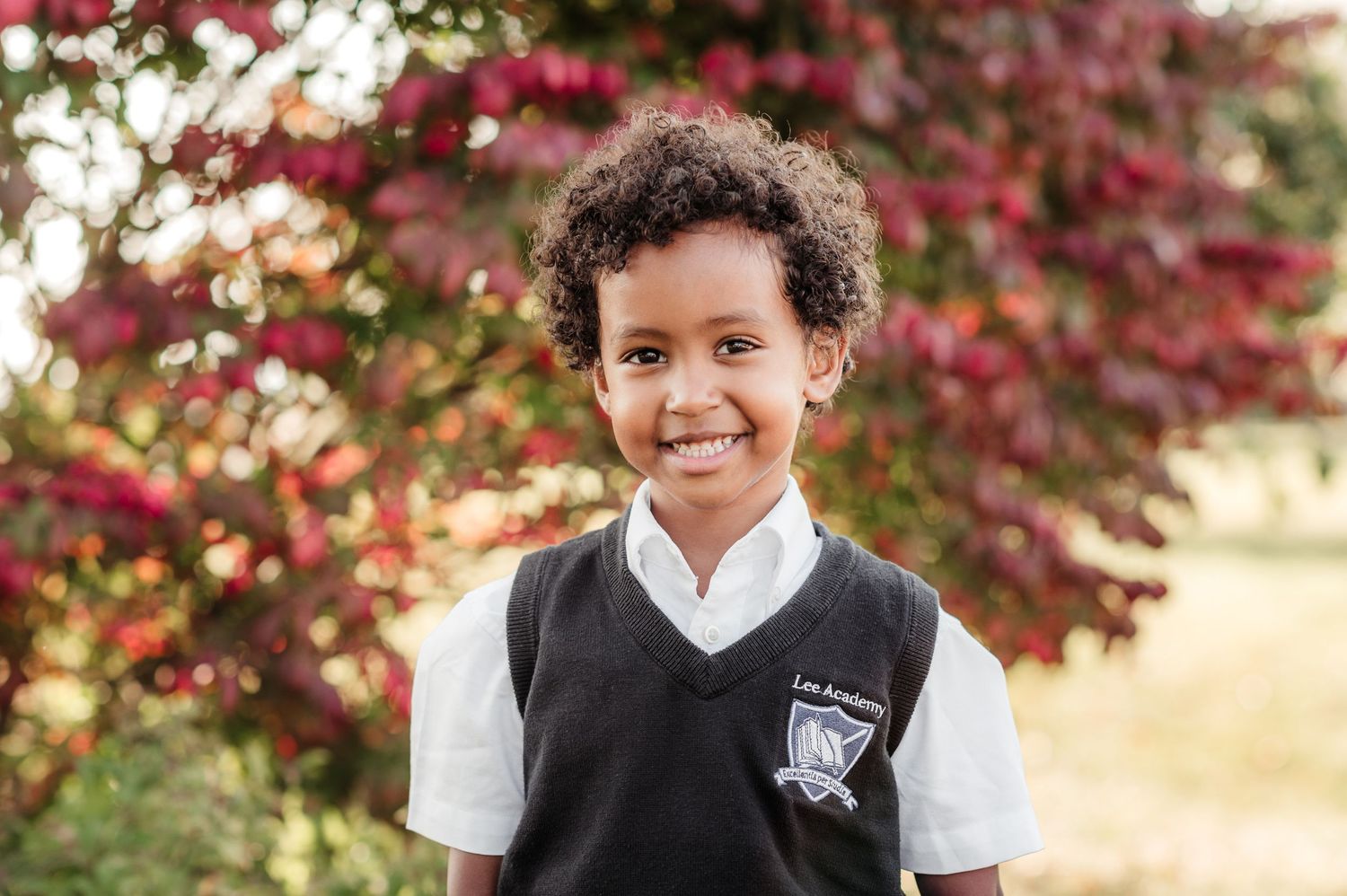 A child in school uniform poses outdoors with vibrant red maple trees in the background during autumn.