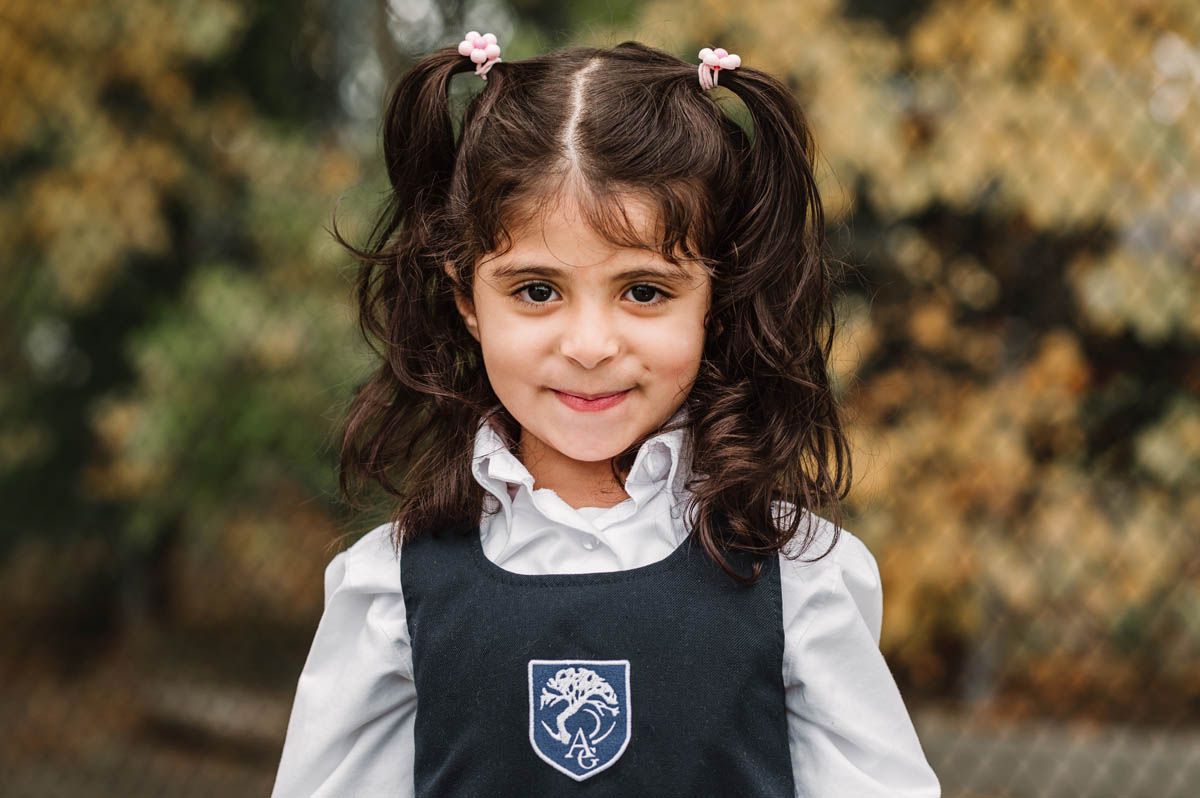 Students in navy and white school uniforms stand outdoors during autumn with chain link fence and foliage in background.
