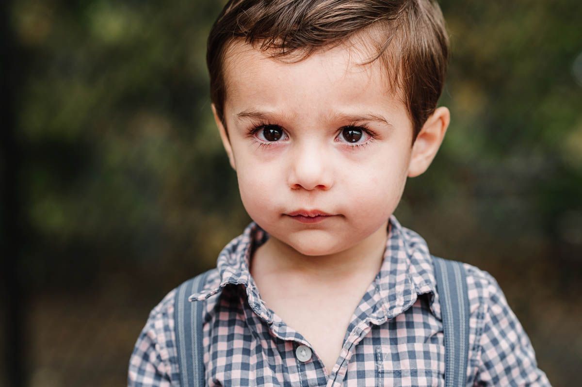 A young child wearing a checkered button-down shirt poses outdoors against a blurred natural background.