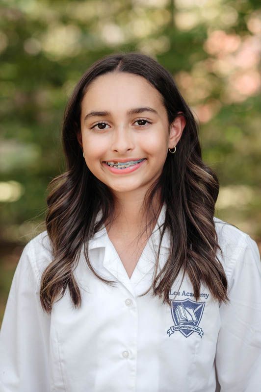 Student in white uniform shirt with embroidered school crest smiles outdoors.