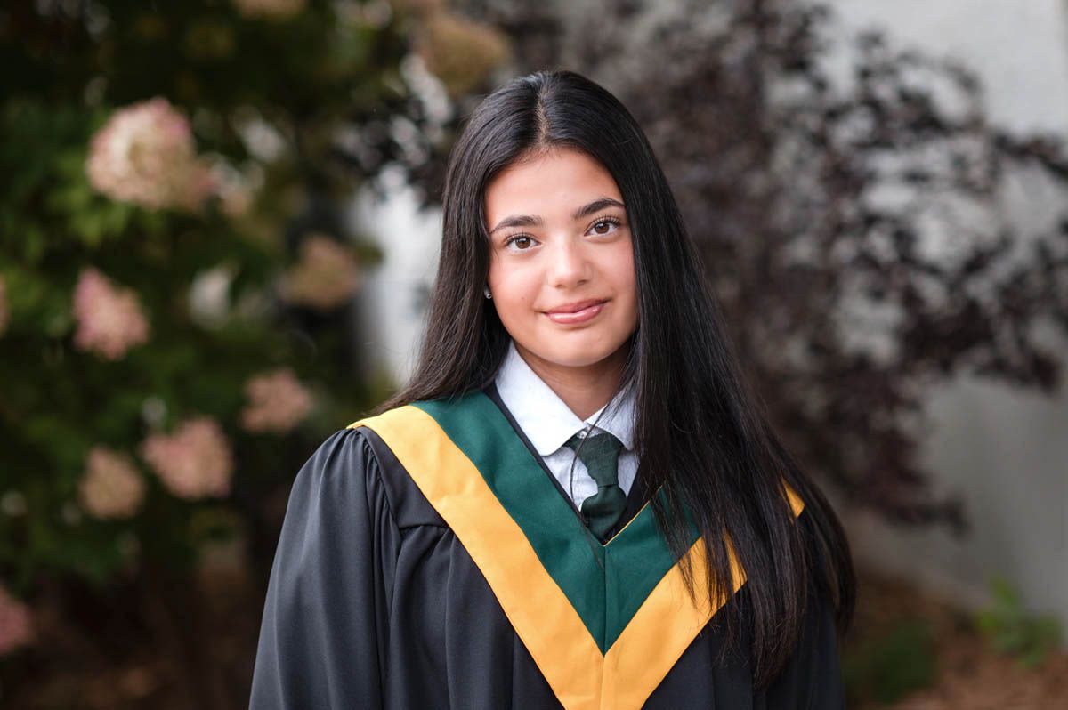 Graduate wearing black gown with yellow sash stands outdoors during ceremony.