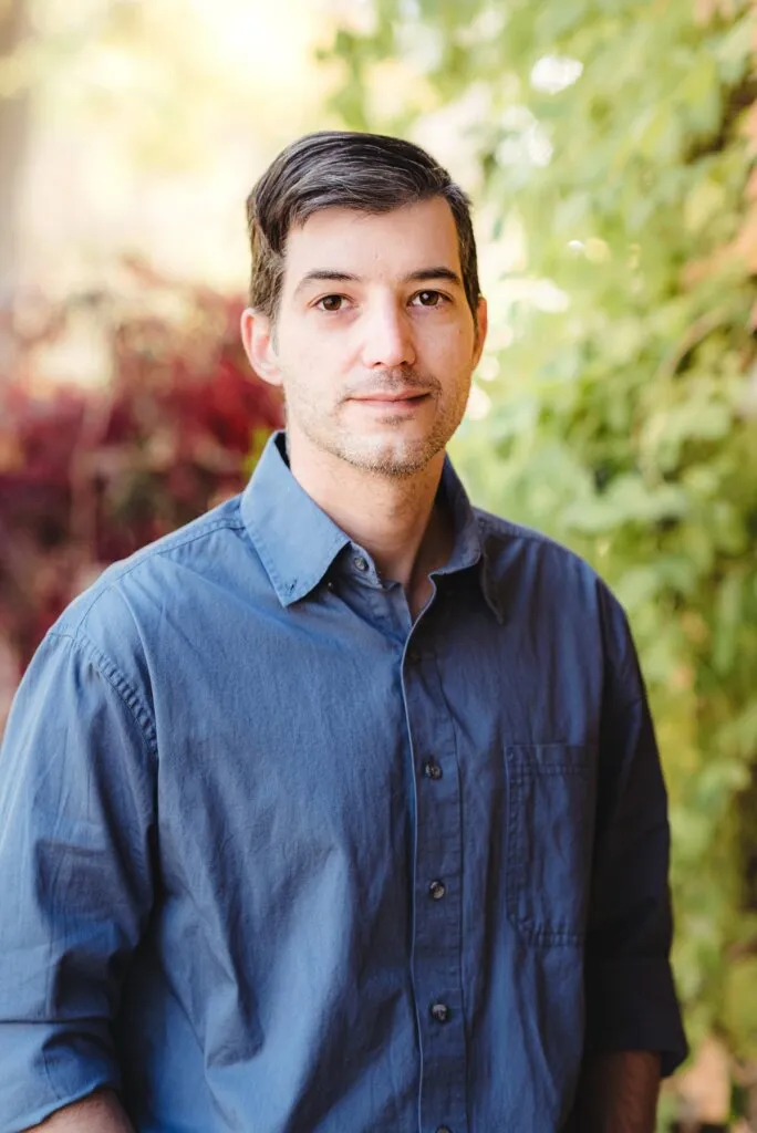 Professional headshot portrait outdoors against blurred natural foliage background wearing a blue button-down shirt.