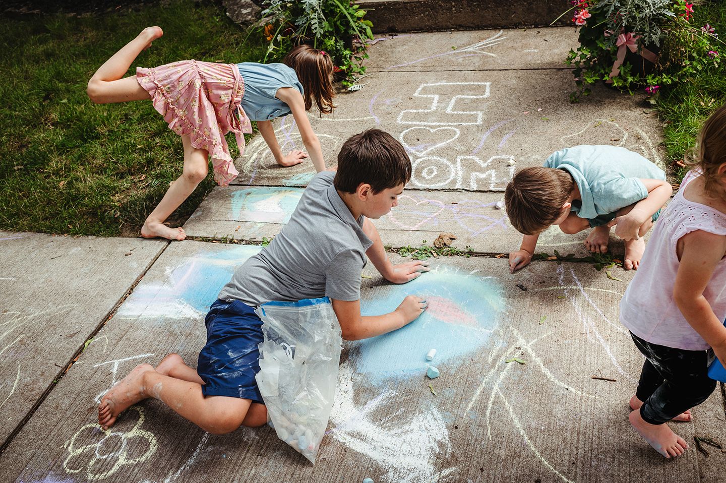 Children drawing with chalk on sidewalk on sunny summer day near flowering bushes.