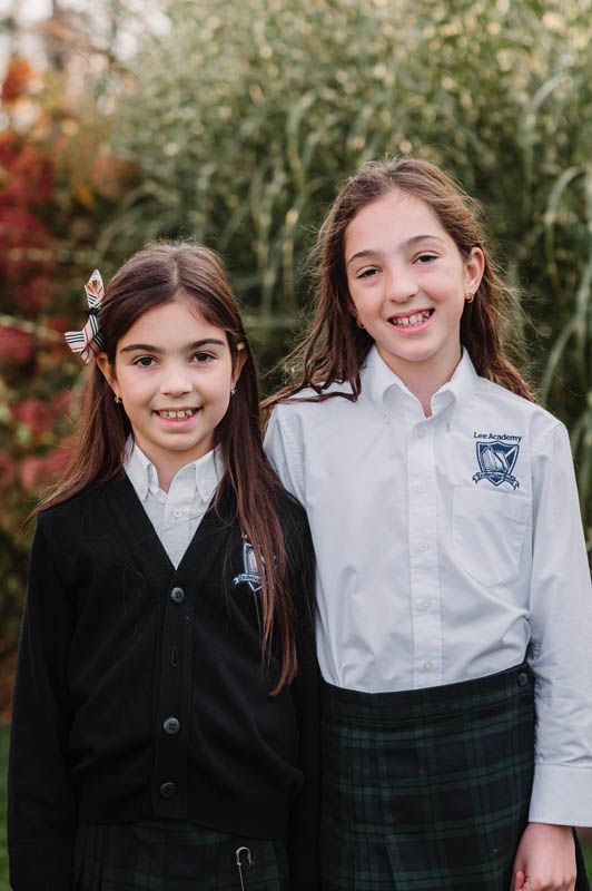 Two students in white shirts and dark school uniforms smile together during outdoor photoshoot.