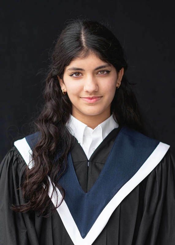 Graduate poses in traditional black graduation gown with white trim against dark background.