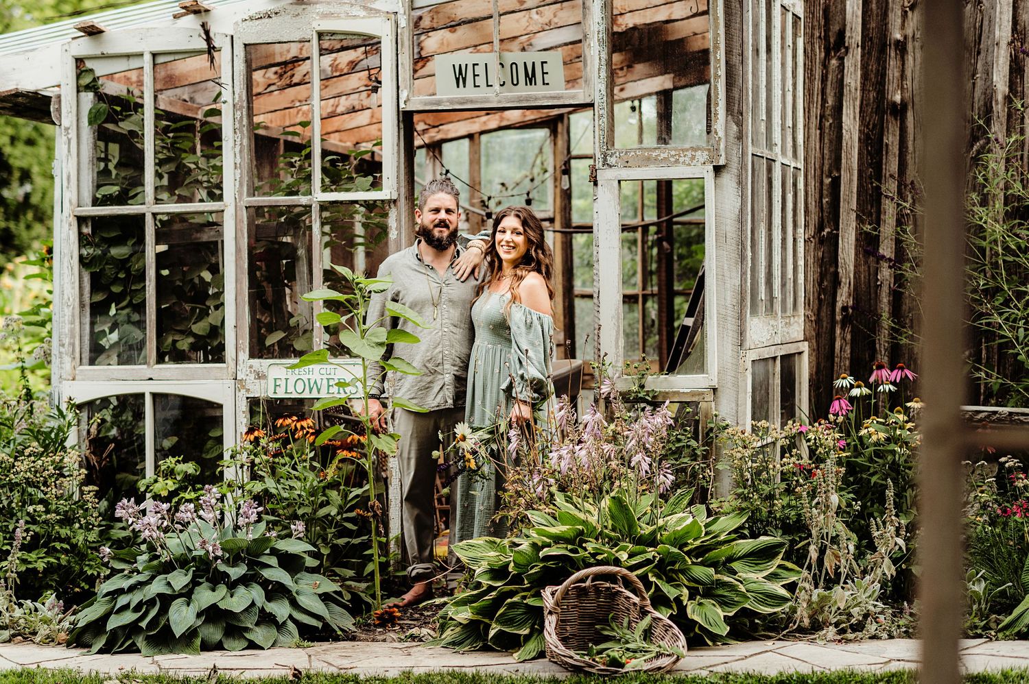 Couple standing in front of rustic greenhouse with plants and windows.