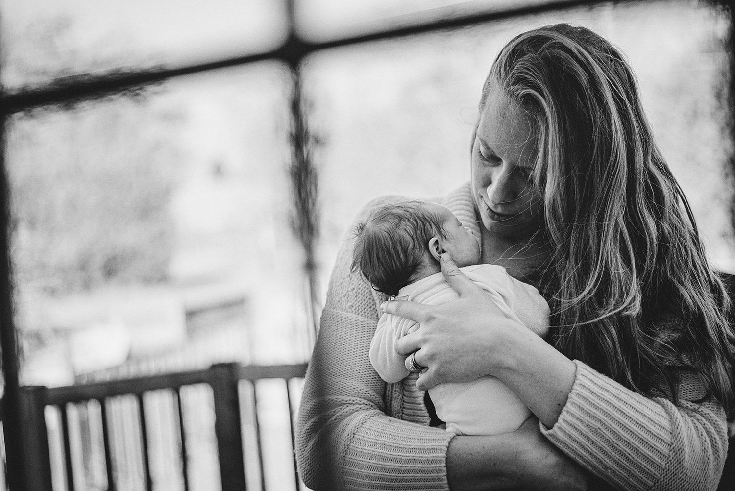 Black and white portrait capturing intimate bonding moment between mother and newborn on outdoor porch.