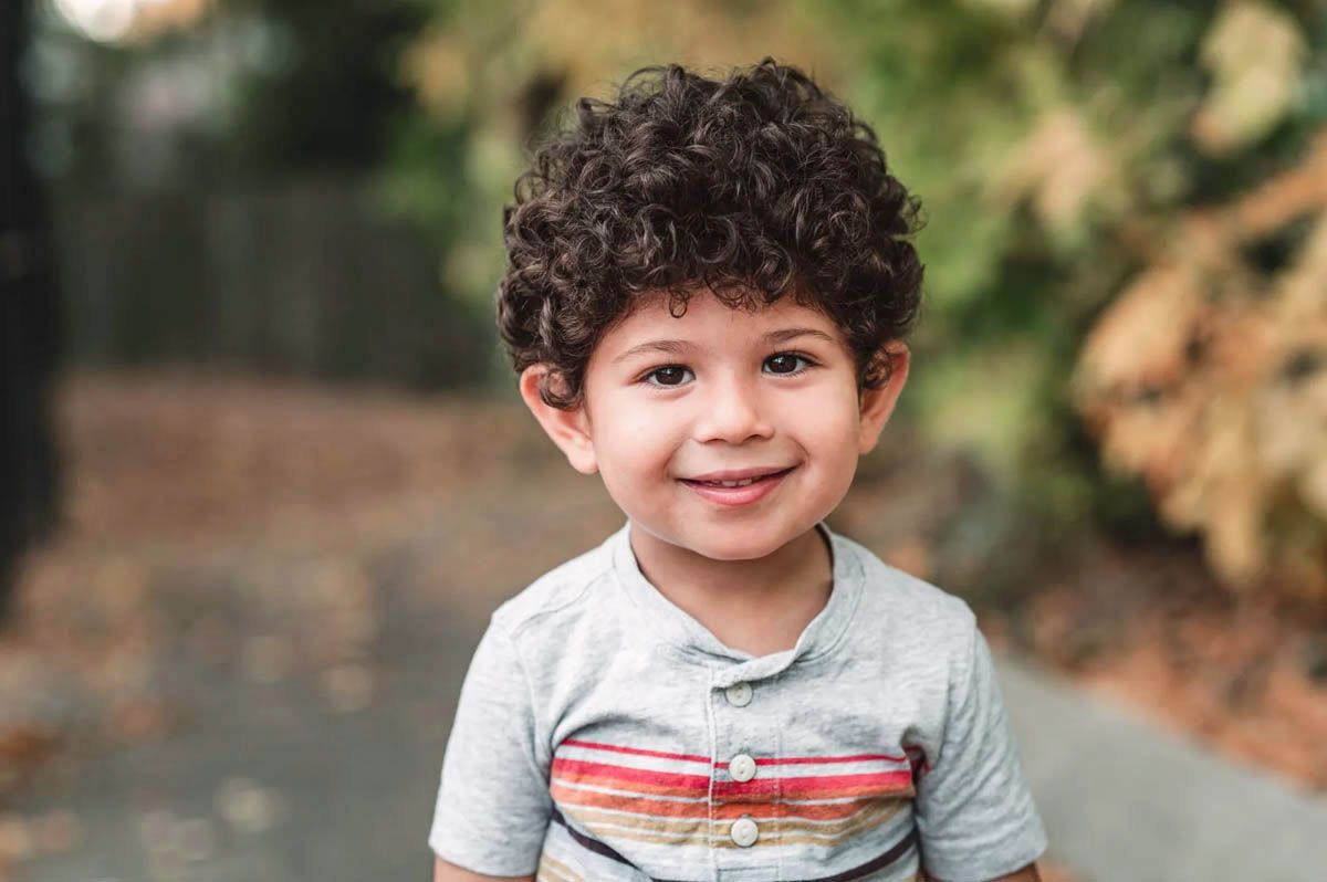Adorable small child wearing a gray hoodie smiles while sitting outdoors in natural lighting.