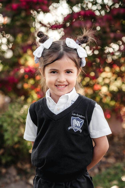 Elementary student in black vest uniform poses cheerfully against colorful garden background.