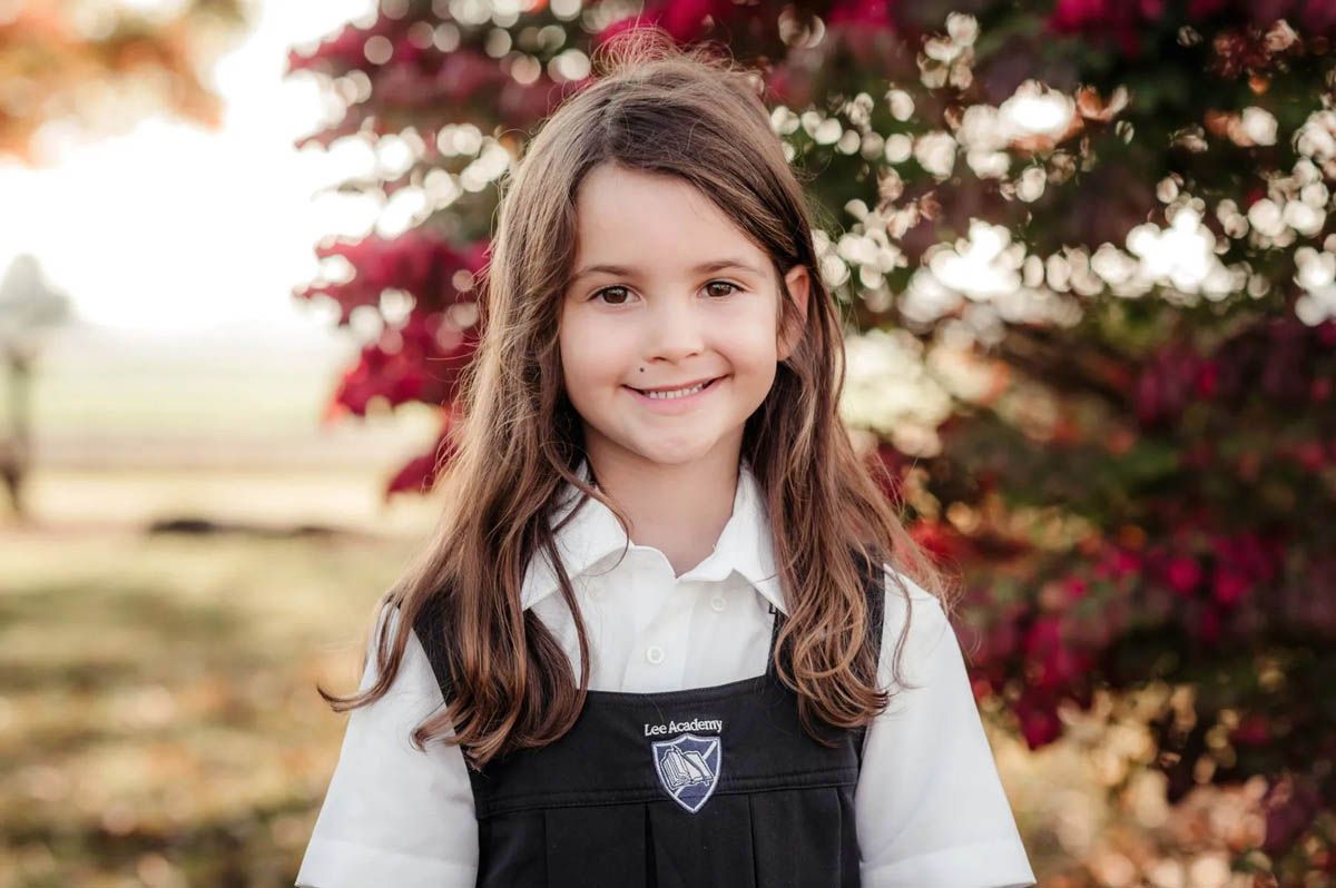 Student in school uniform smiles brightly while posing outdoors with red flowering bushes in background.