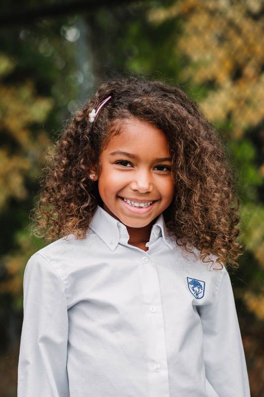 A student in white school uniform shirt with crest smiles brightly against autumn foliage background.