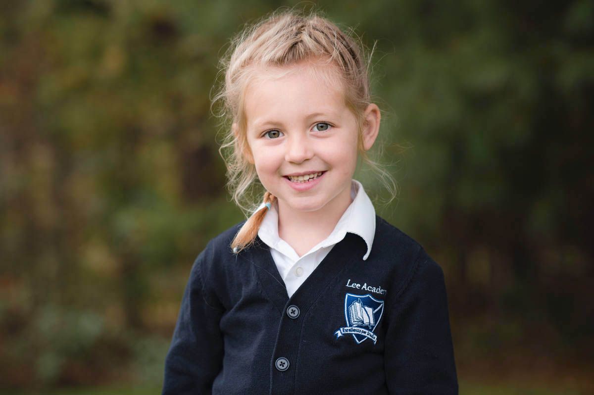 A student wearing a navy blue uniform sweater smiles while posing for a school portrait outdoors.