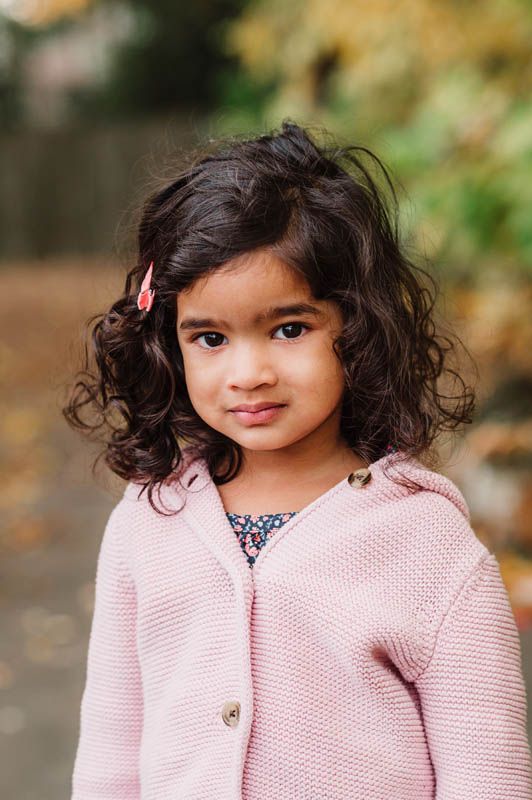 Young child wearing a pink knit sweater poses outdoors with curly dark hair during an autumn photoshoot.