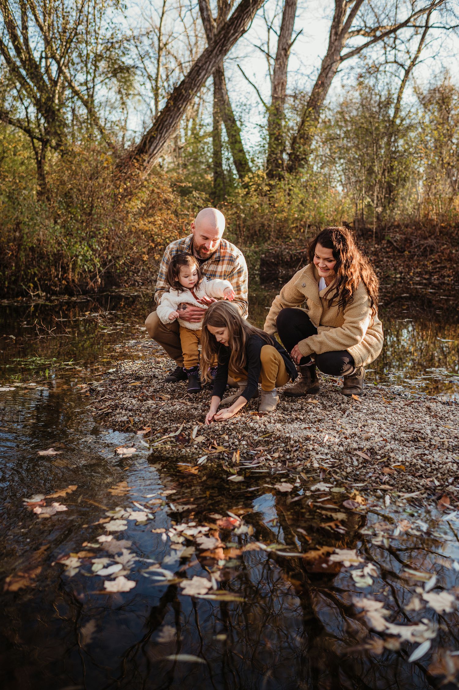 Family exploring creek bed with fallen leaves during golden hour in wooded area.