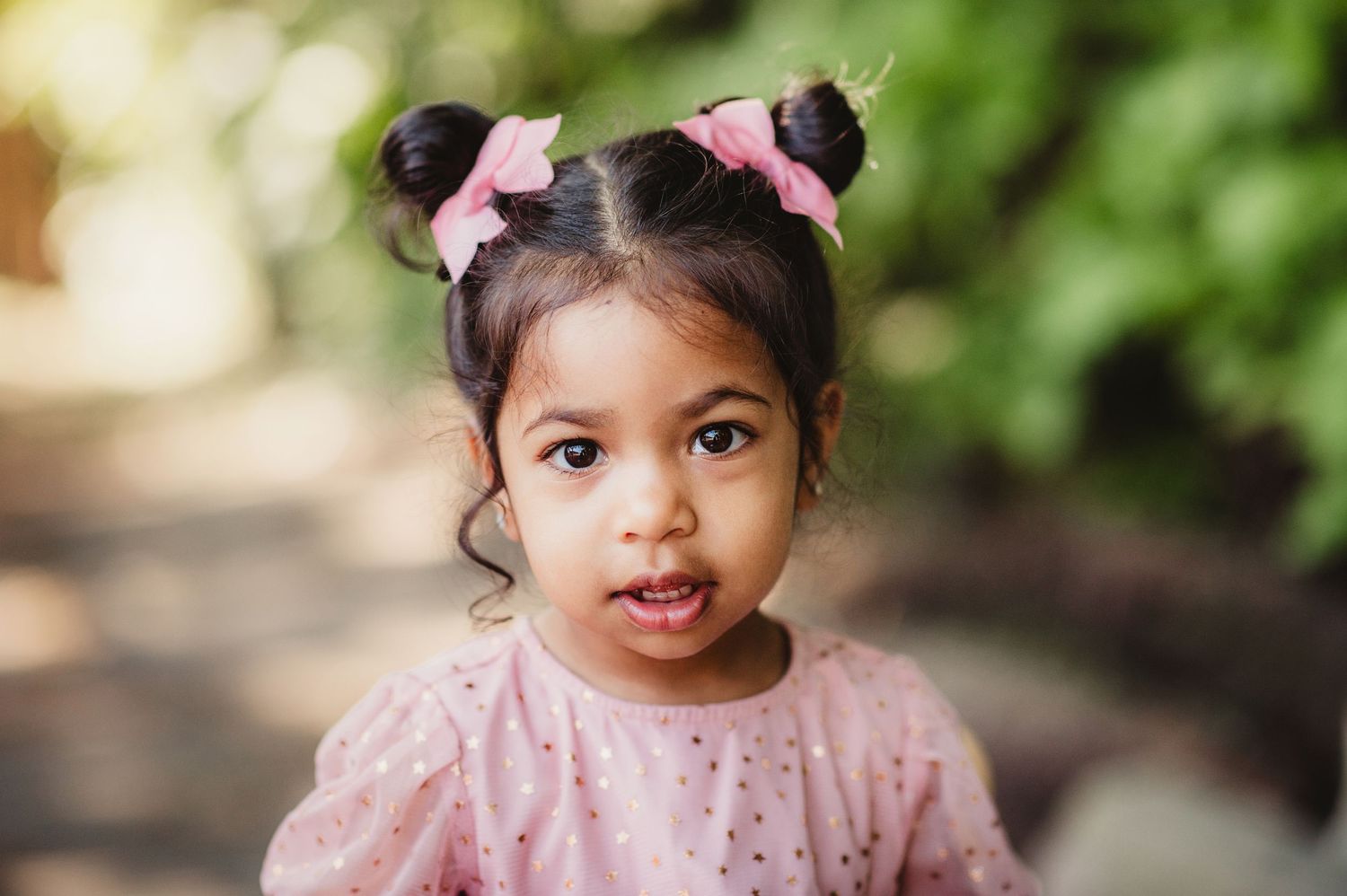 A child in a pink dress with matching hair accessories sits amid outdoor greenery.