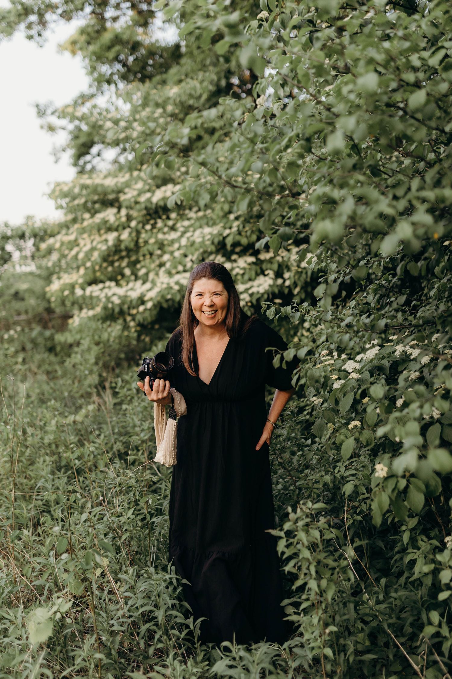 Woman in black dress poses joyfully in front of lush green foliage.
